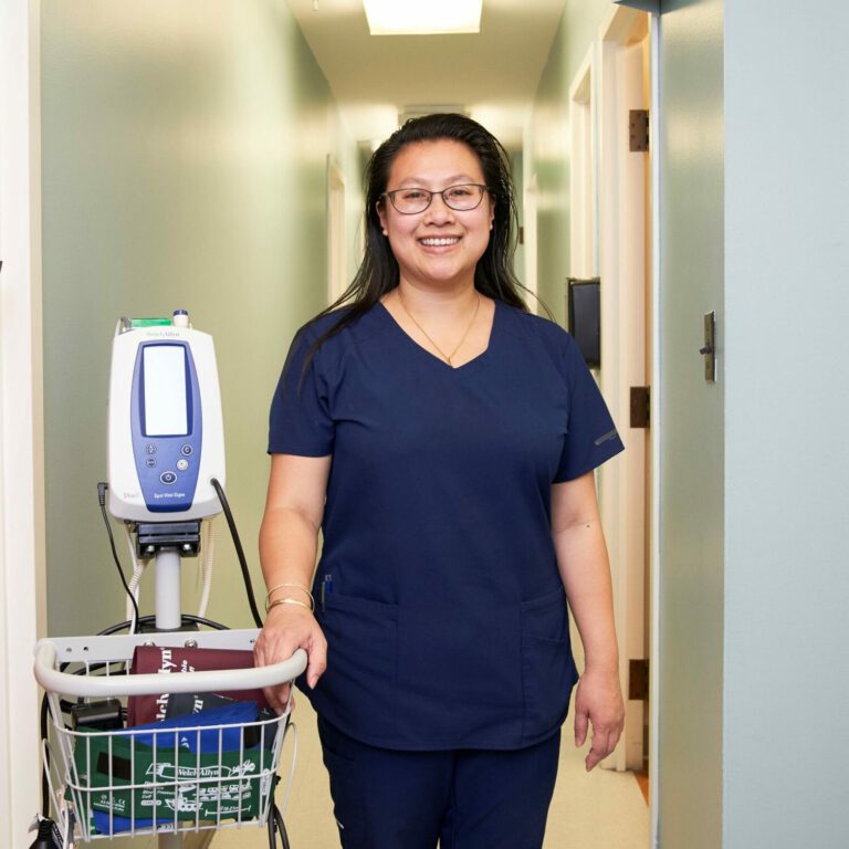 Nurse standing in hallway next to blood pressure machine.